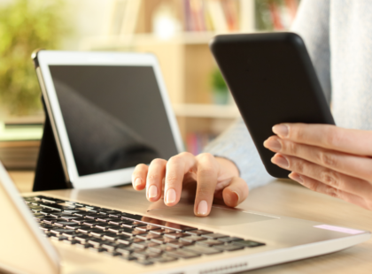 An image of a woman's hands holding a smart phone in one and on a laptop keyboard in the other with an ipad open on the desk in the background